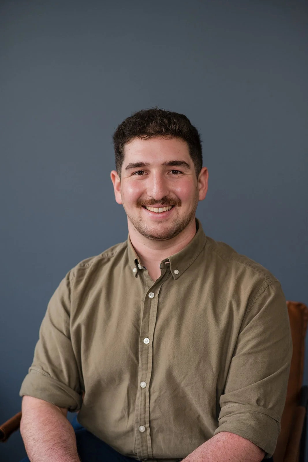 A young man with short dark hair and a beard, smiling, sitting against a plain blue background, wearing a khaki button-up shirt with the sleeves rolled up.