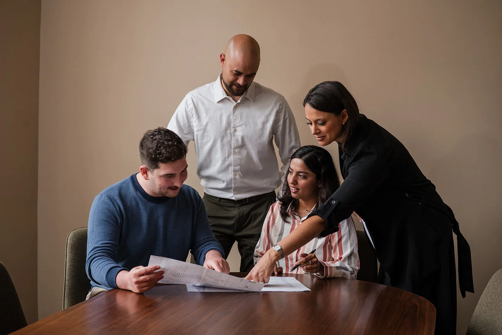 A diverse group of five people in business casual clothing engaged in a discussion around a table, examining documents, with one woman pointing at something on the papers.