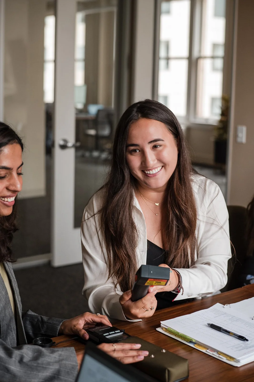 Two women smiling at a meeting, one woman holds a card reader, and there are notebooks and papers on the table.
