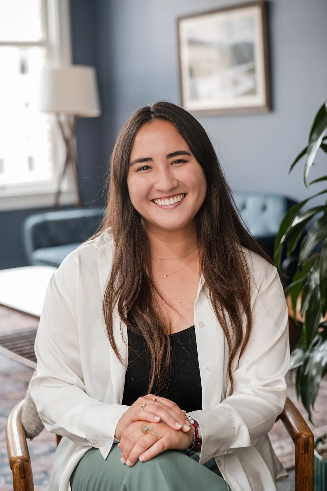 A smiling woman with long dark hair, wearing a white shirt and sitting in a room with a blue wall, framed picture, green plant, and window.
