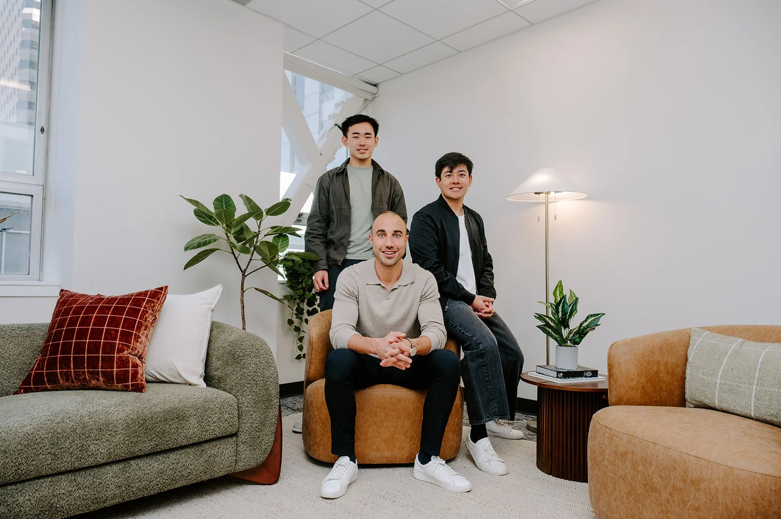 Three men in a modern living room with green and beige sofas, a floor lamp, and potted plants, posing for a photo.
