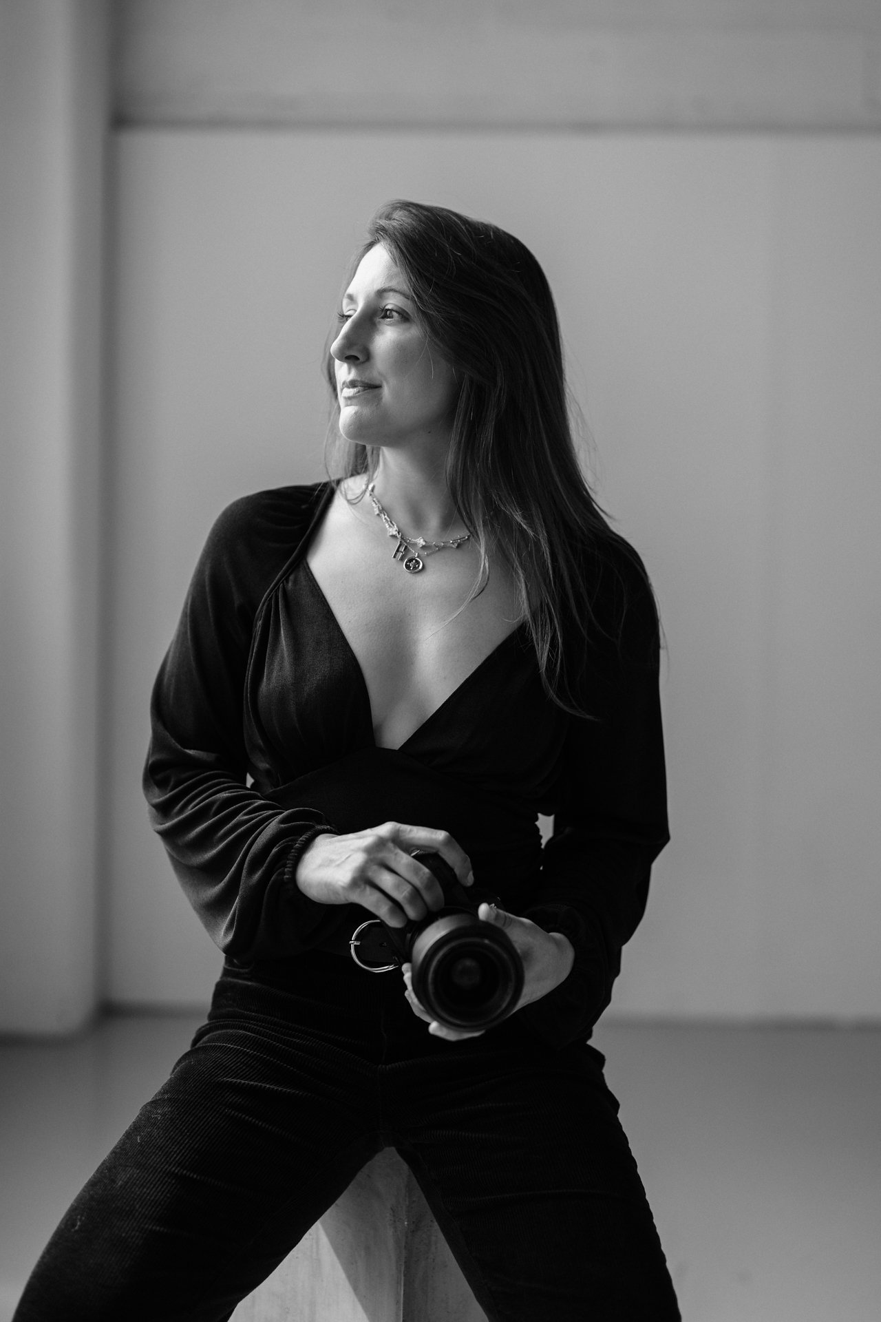 A woman with dark hair, wearing a black top and necklaces, holding a camera, standing in front of a plain wall.