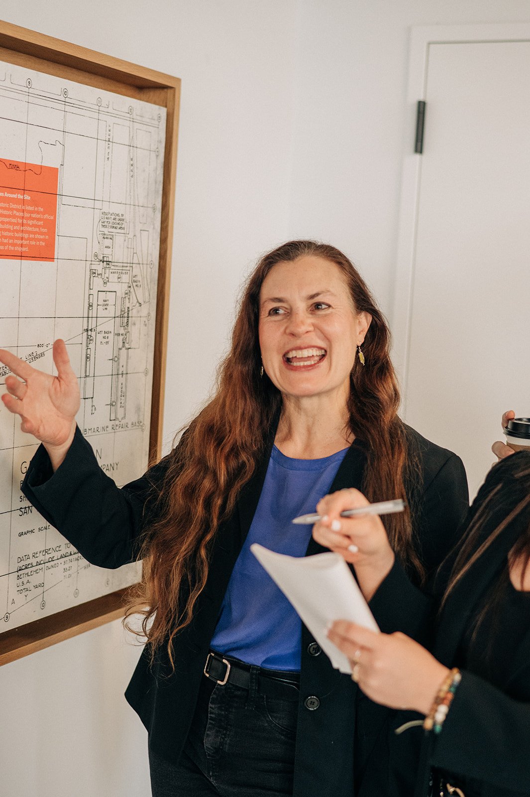 A woman with long, wavy red hair gesturing and speaking while holding a pen and notepad, standing next to a person holding a coffee cup, in front of a large blueprint on a wall.