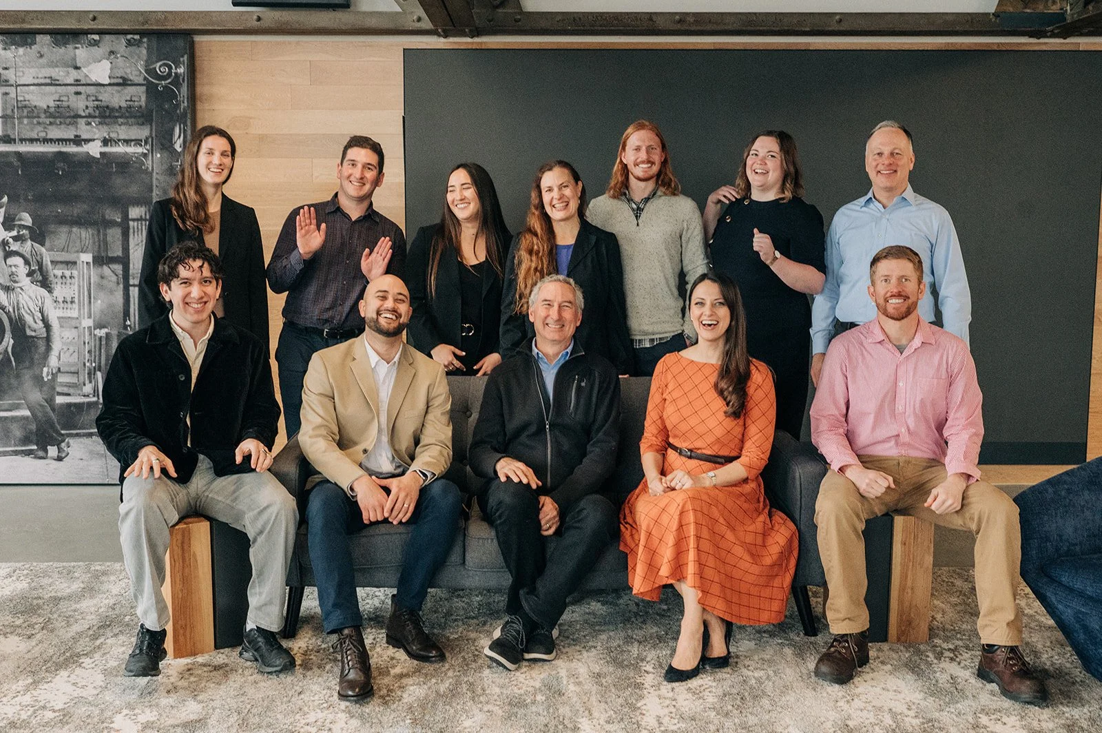 A group photo of fifteen diverse individuals, including men and women, smiling and standing or sitting together in a modern office space with wood and black wall accents, and a black-and-white historical photograph on the wall.