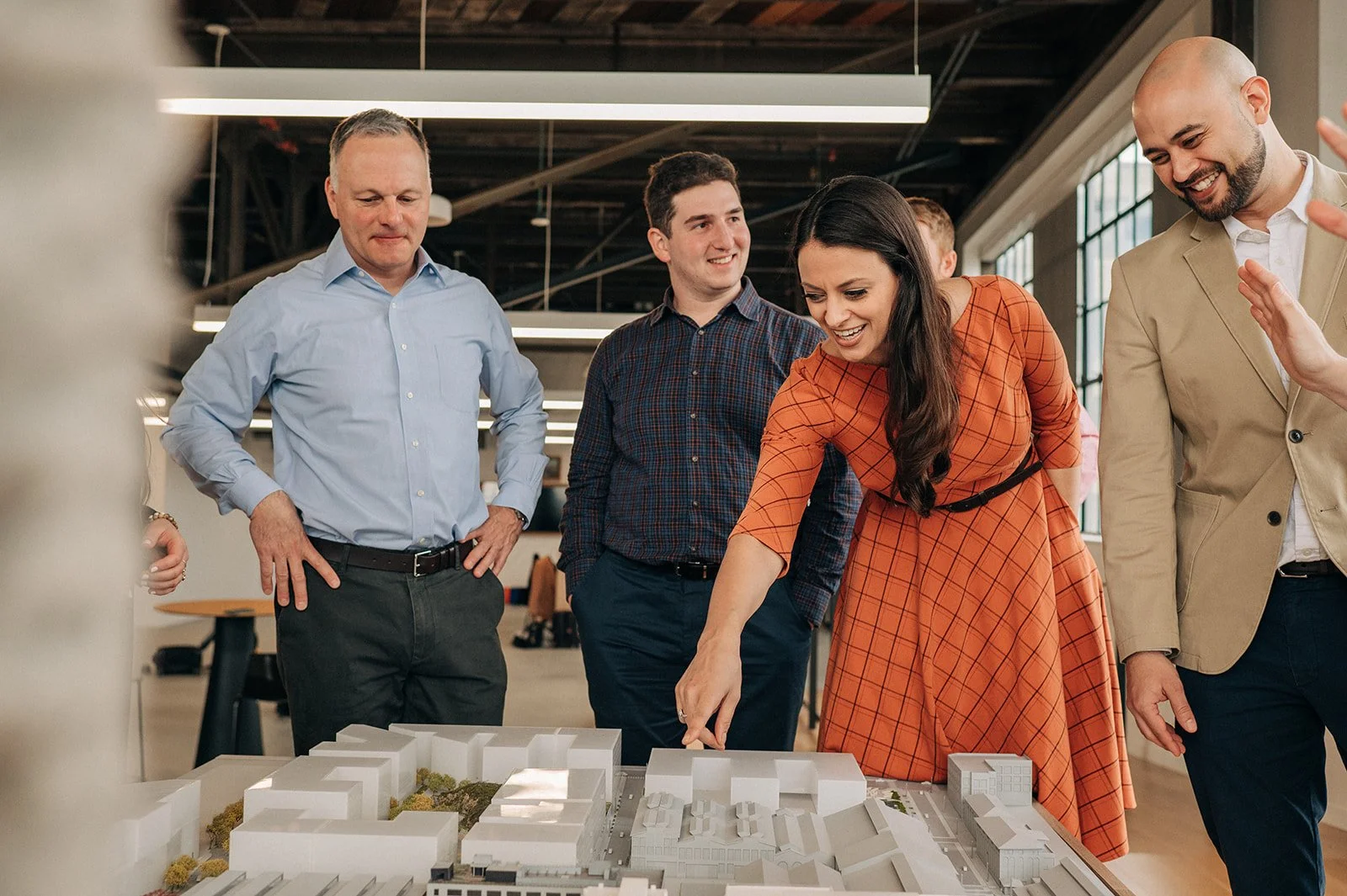 A group of five diverse professionals, including a woman in an orange dress, gathered around an architectural model on a table in a modern office space with large windows and high ceilings, discussing and pointing at the model.