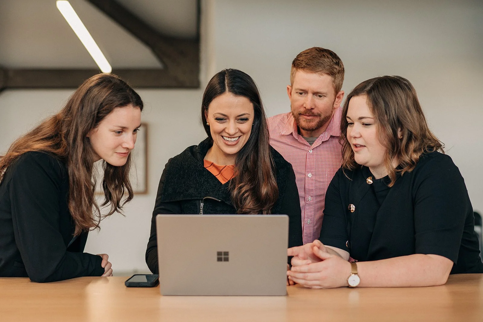 Four people looking at a laptop together, smiling and engaged, in a modern indoor setting.