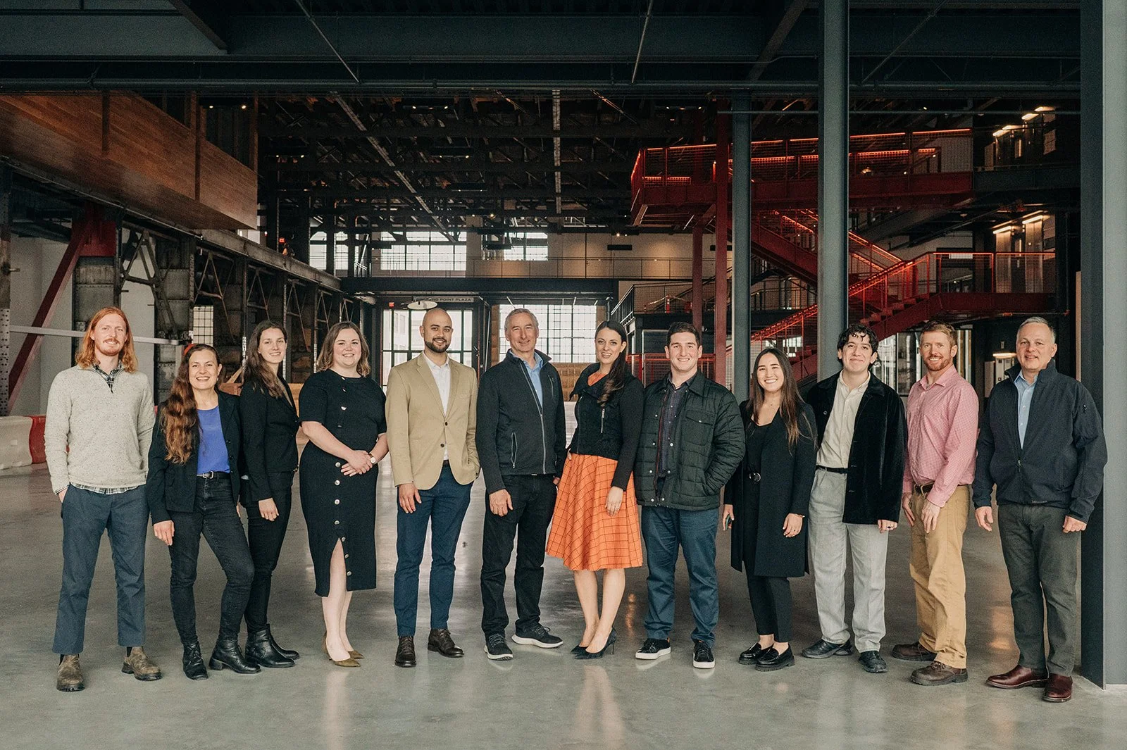 Group of fifteen diverse people standing in a modern industrial-style building with red staircase and large windows