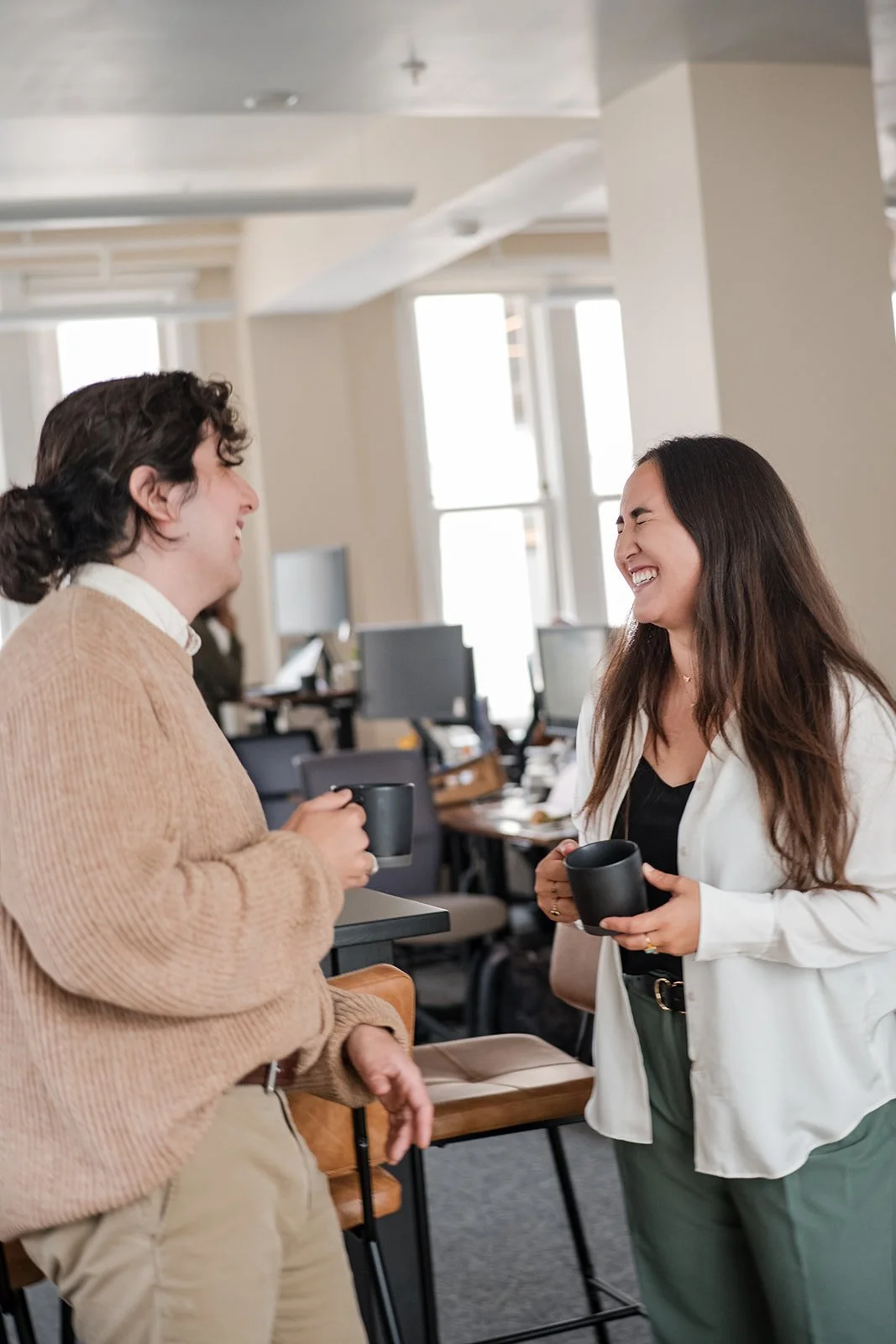 Two women with dark hair are smiling and holding black mugs, having a friendly conversation in an office environment with desks and computers.