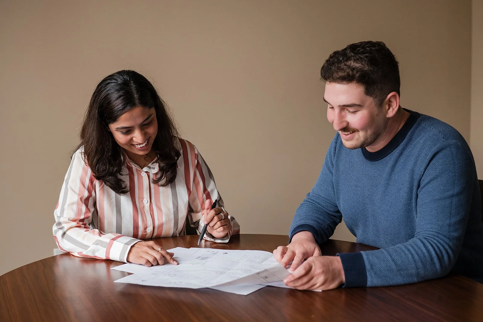 A woman and a man sitting at a round table, smiling and looking at documents and papers, with the woman holding a pen.