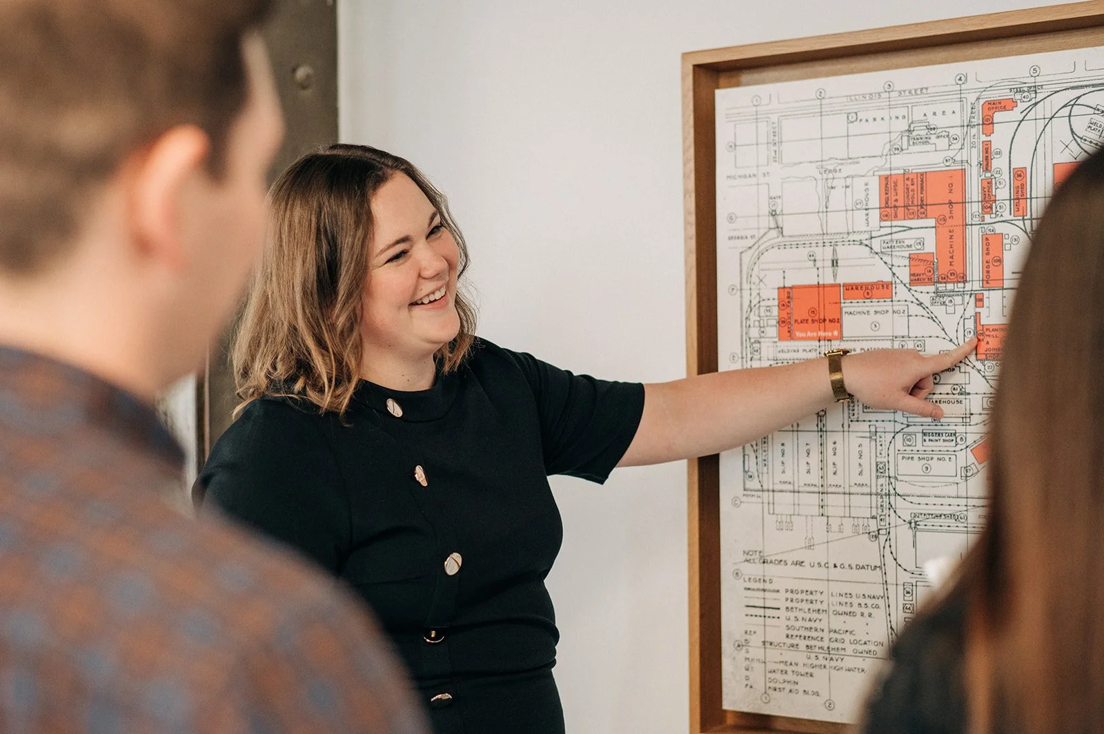 A woman with shoulder-length brown hair, wearing a black dress, points at a detailed blueprint or site plan on a wall, while talking to a group of people in an indoor setting.