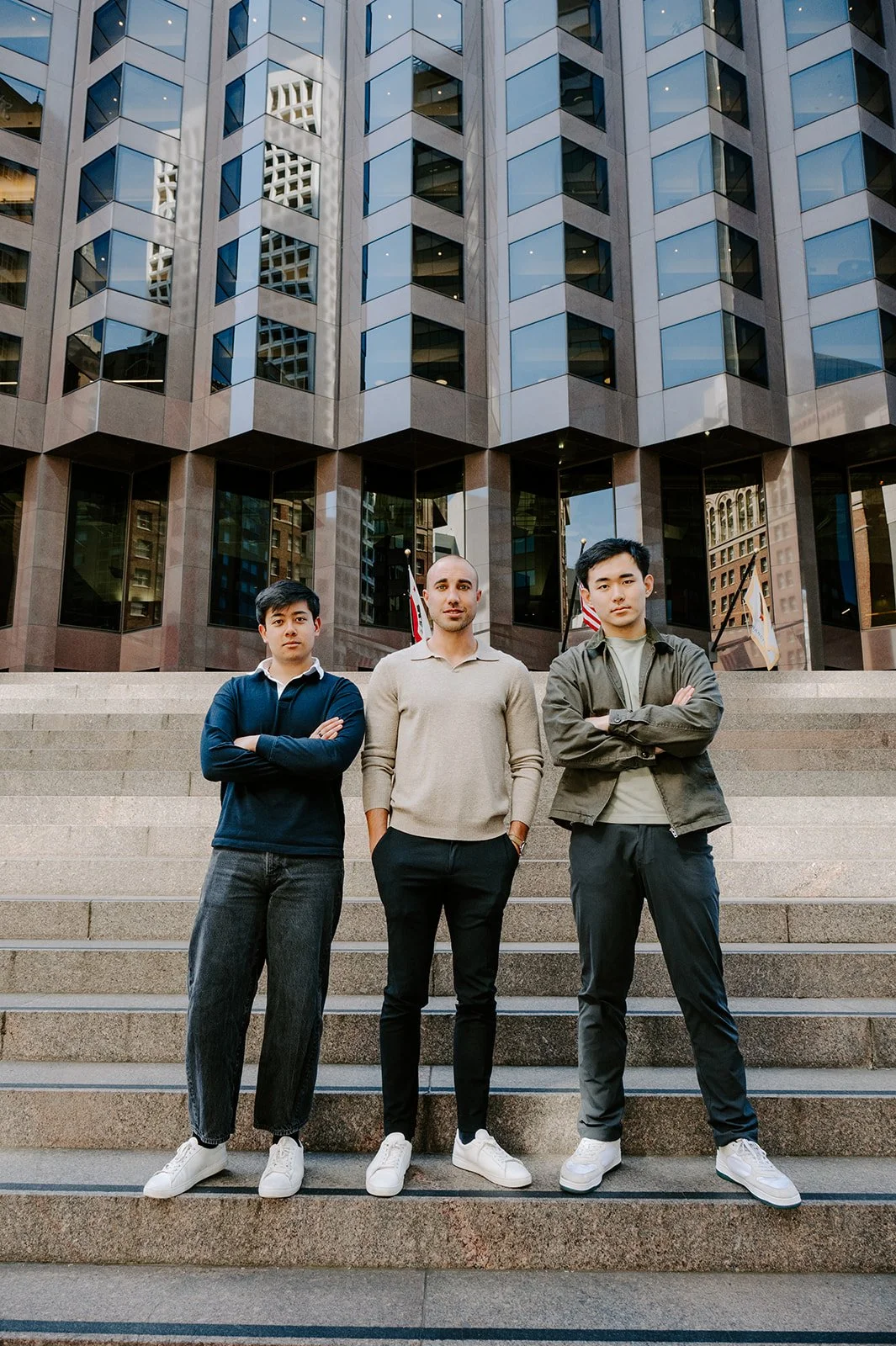 Three young men standing on steps in front of a modern glass office building with flags behind them.
