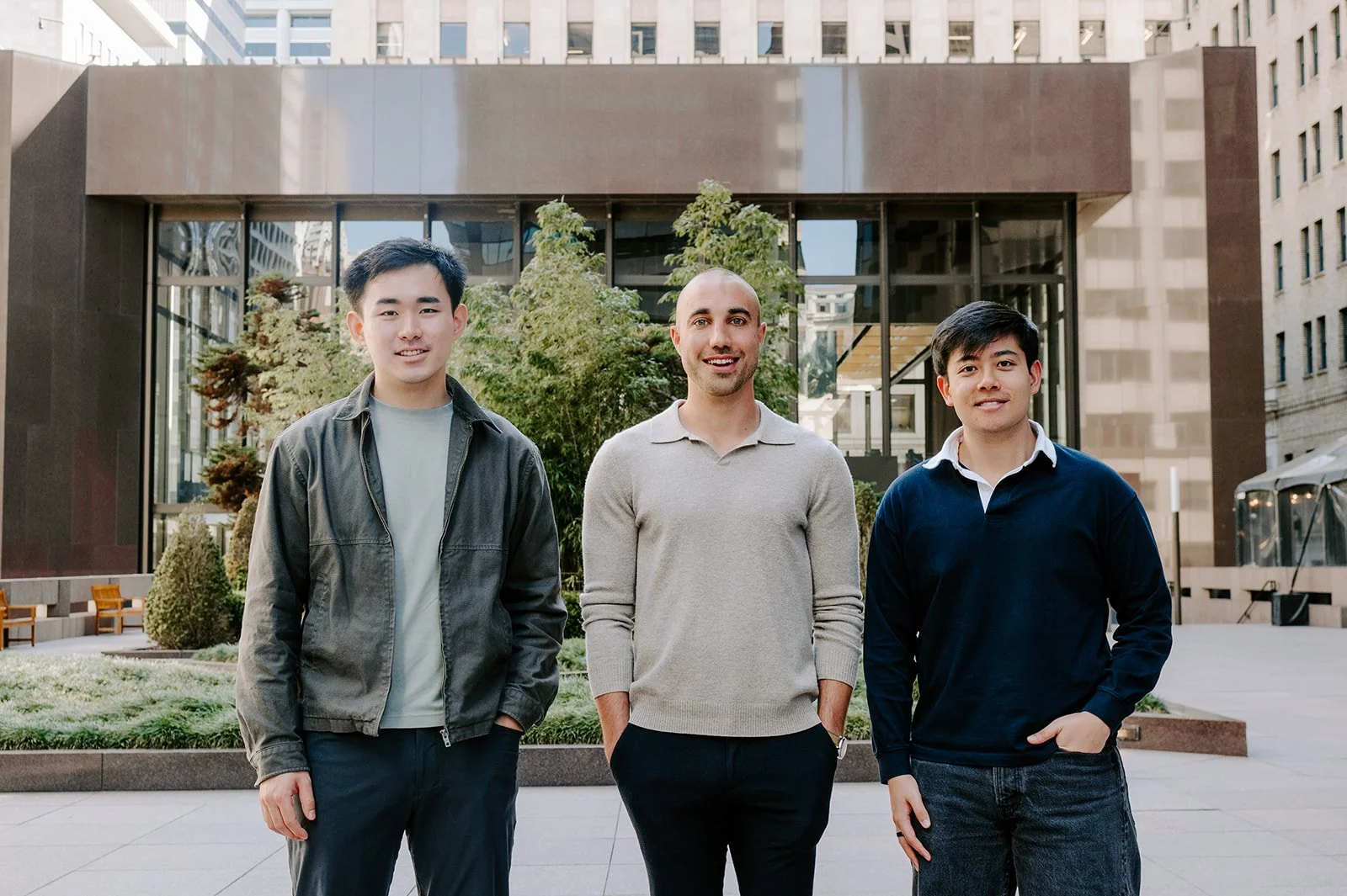 Three men standing outdoors in front of a modern building and trees, smiling at the camera.
