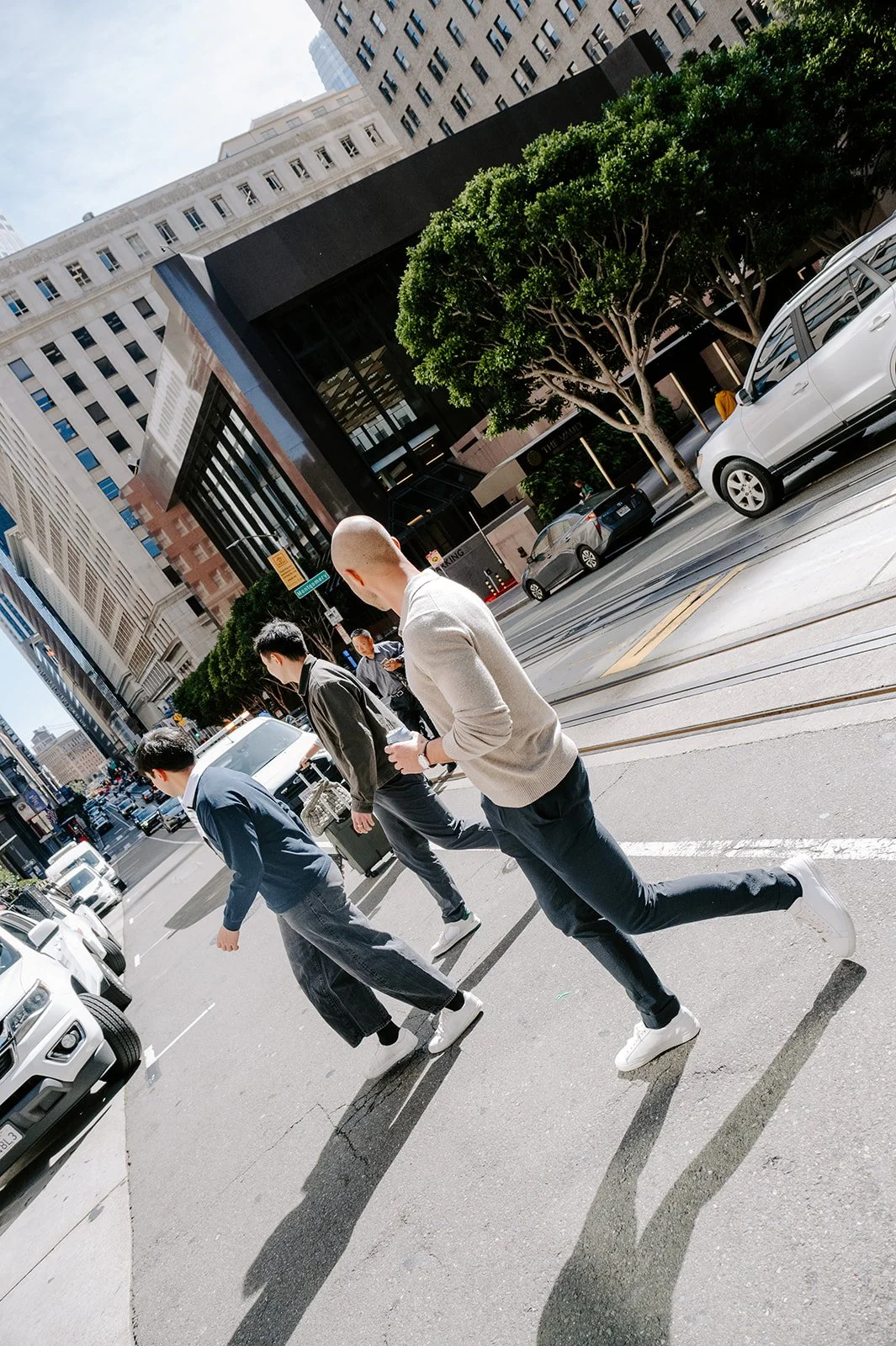 A city street scene with four men crossing at a crosswalk, surrounded by tall buildings, trees, cars, and tram tracks on a sunny day.