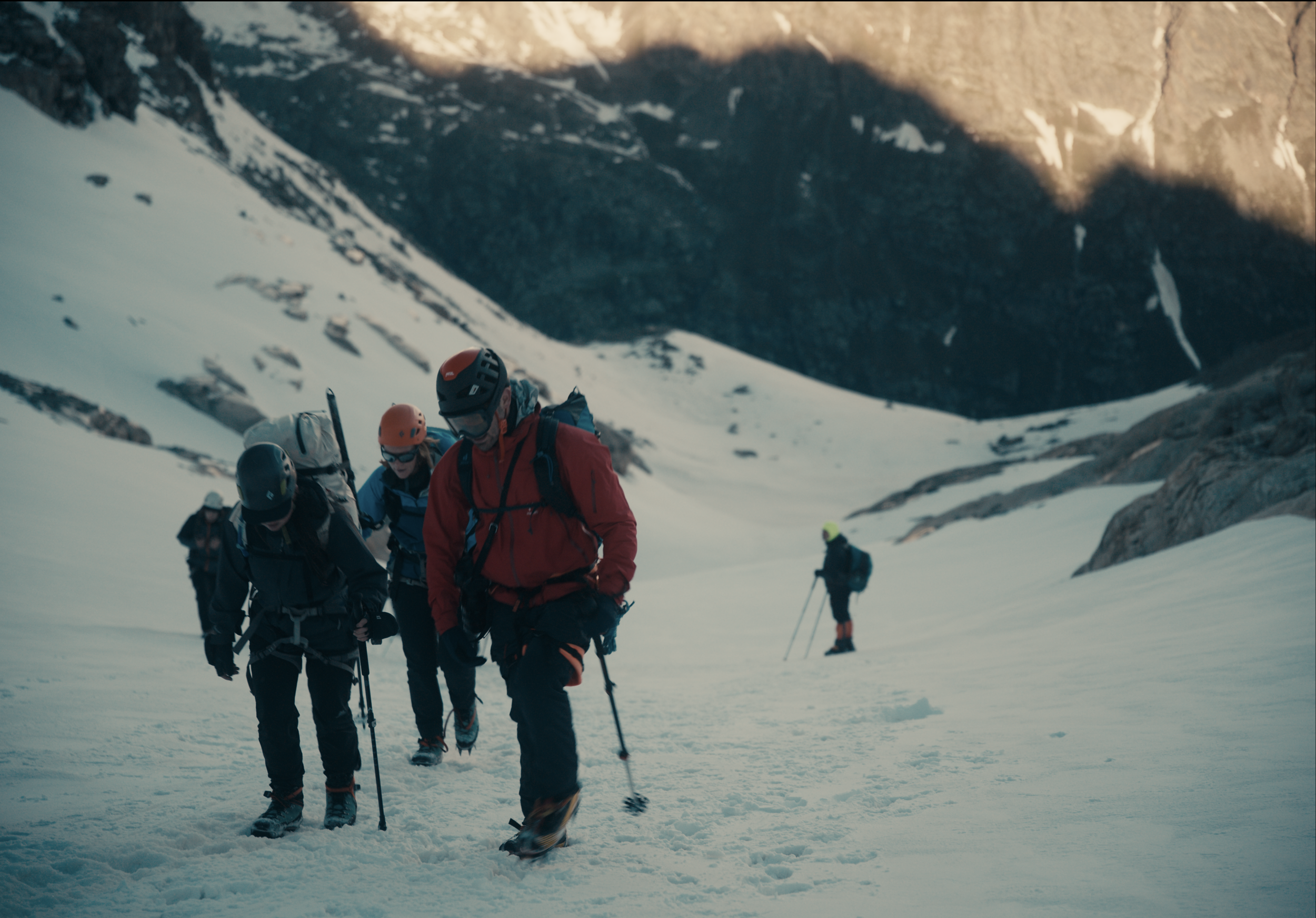 Group of people climbing snow-covered mountain with mountain peaks in background, wearing helmets and outdoor mountaineering gear.