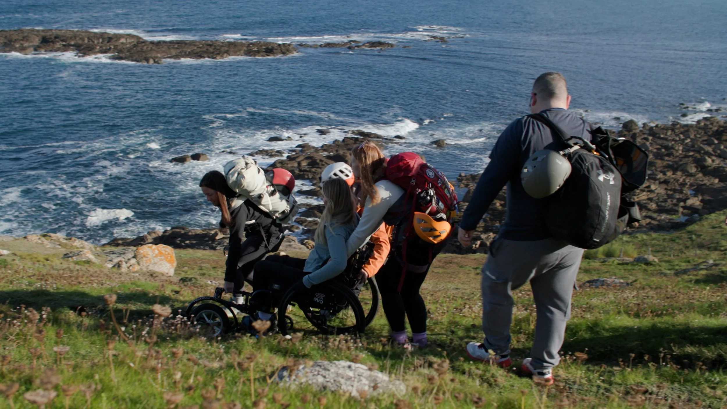 Four people navigating rocky coastal terrain, including a wheelchair user, wearing backpacks and helmets with the sea visible in the background."