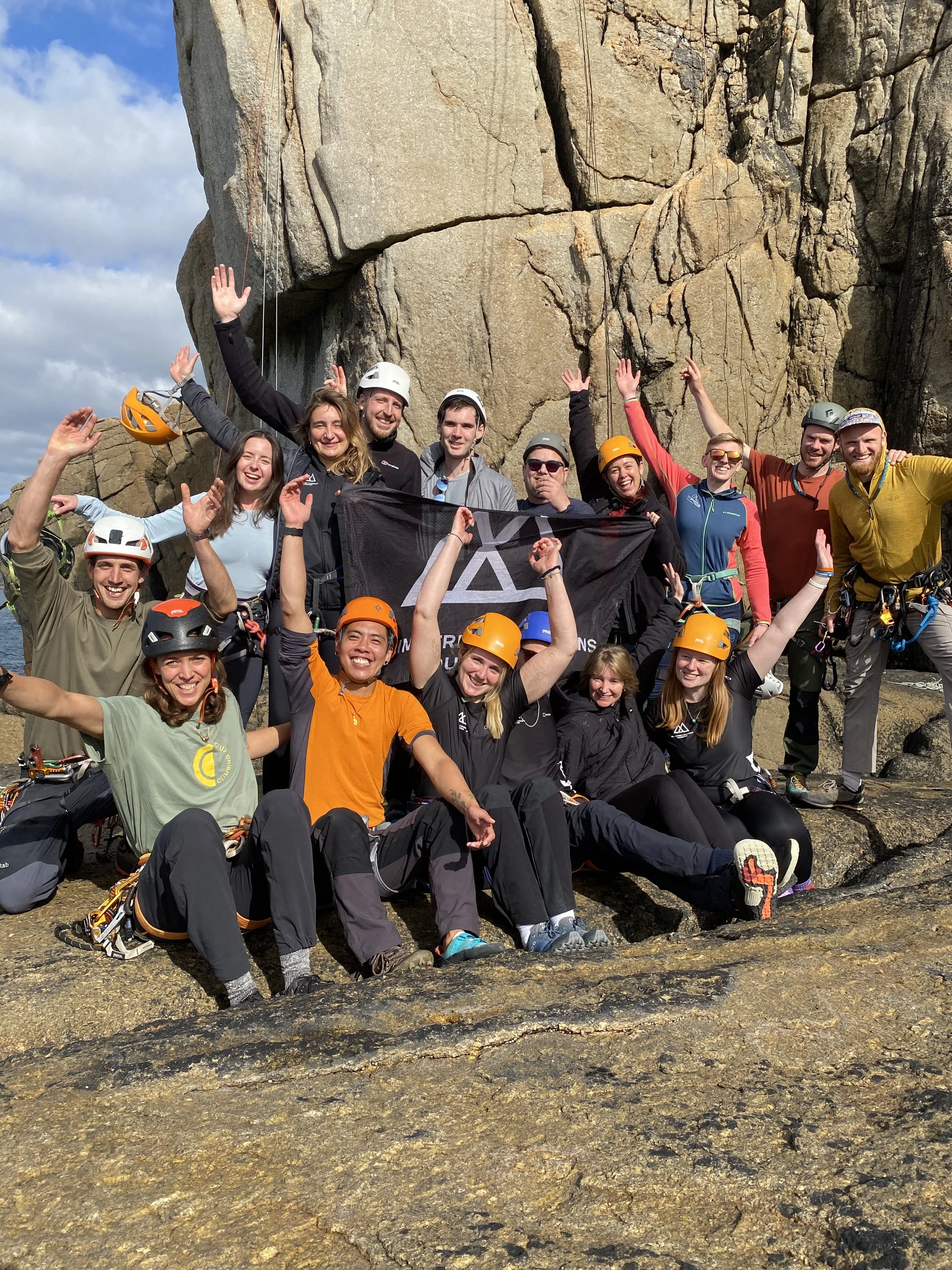 Group of rock climbers celebrating at the base of a large rock face, some holding a black flag with white logo, all wearing helmets and outdoor climbing gear.