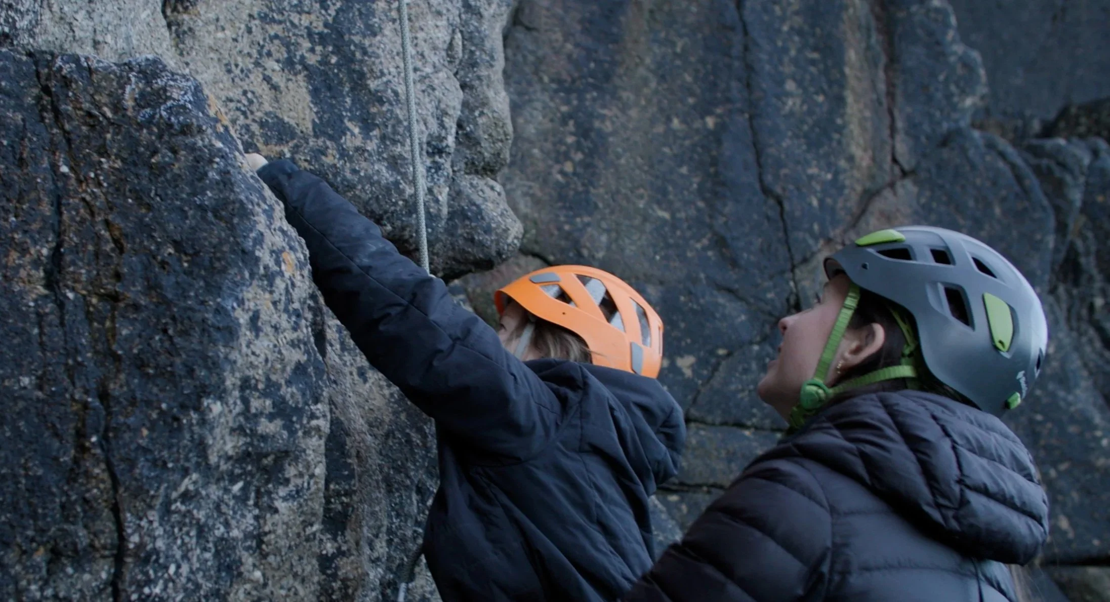 Two people wearing helmets and jackets climbing a rocky wall outdoors.