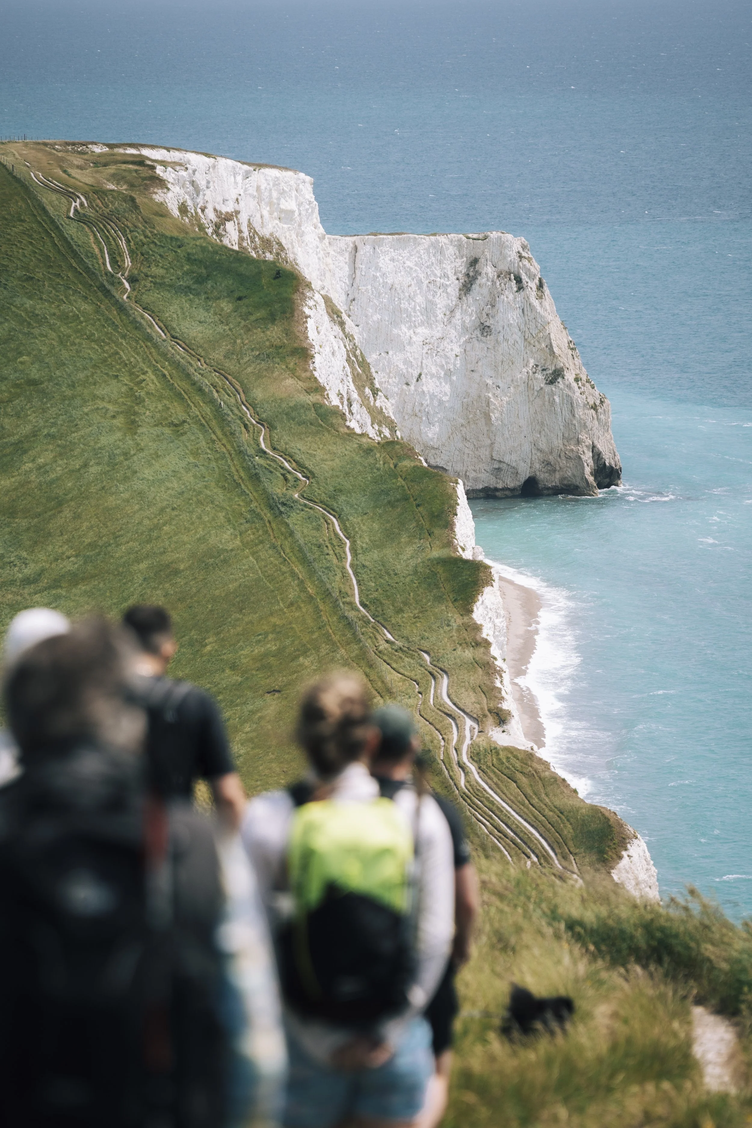 People walking along a grassy trail on a cliffside overlooking the ocean, with white chalk cliffs and blue water in the background.