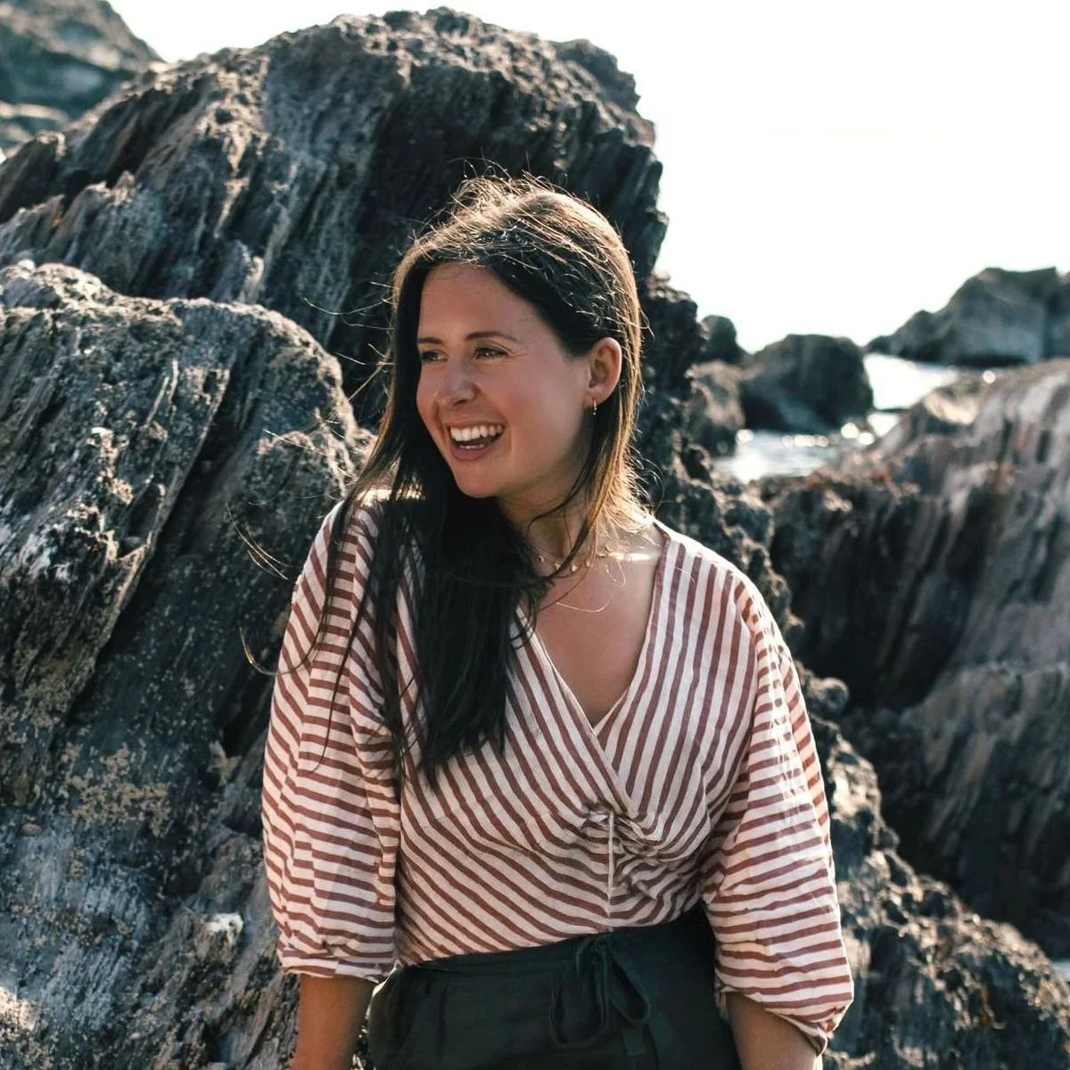 A 30-something year old woman with long dark hair smiling and looking to her right, wearing a striped top, standing near rocky cliffs by the sea.