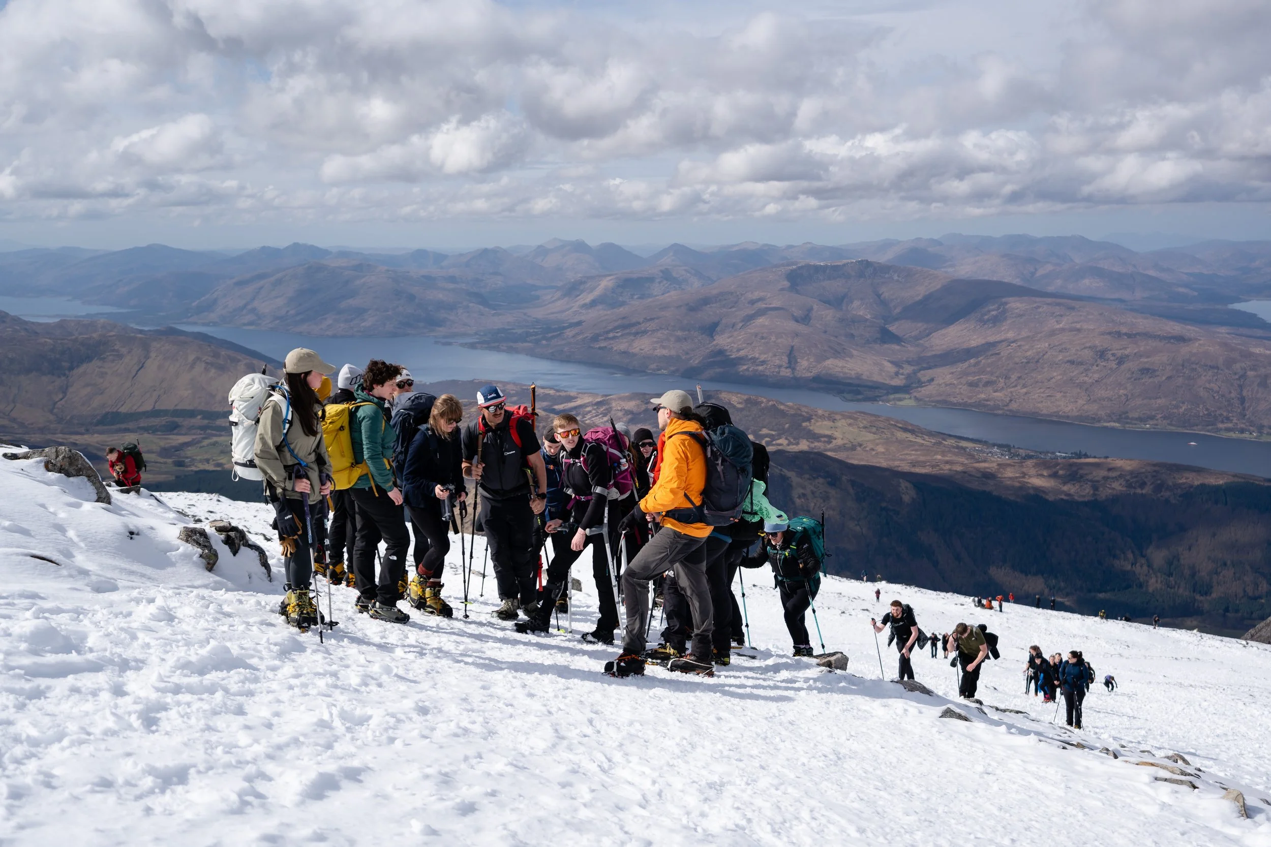 Group of hikers in winter gear ascending a snowy mountain trail, with scenic mountains and lakes in the background, under cloudy sky.