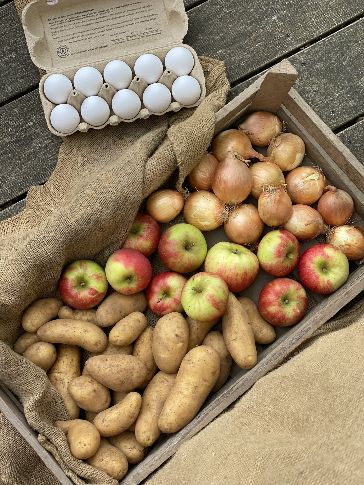 Karton mit weißen Eiern, Zwiebeln, Kartoffeln und Äpfeln in einem Holzkasten auf einer Holztisch. Zeigt Sortiment aus dem Hofladen von Kartoffelhof Schäffer