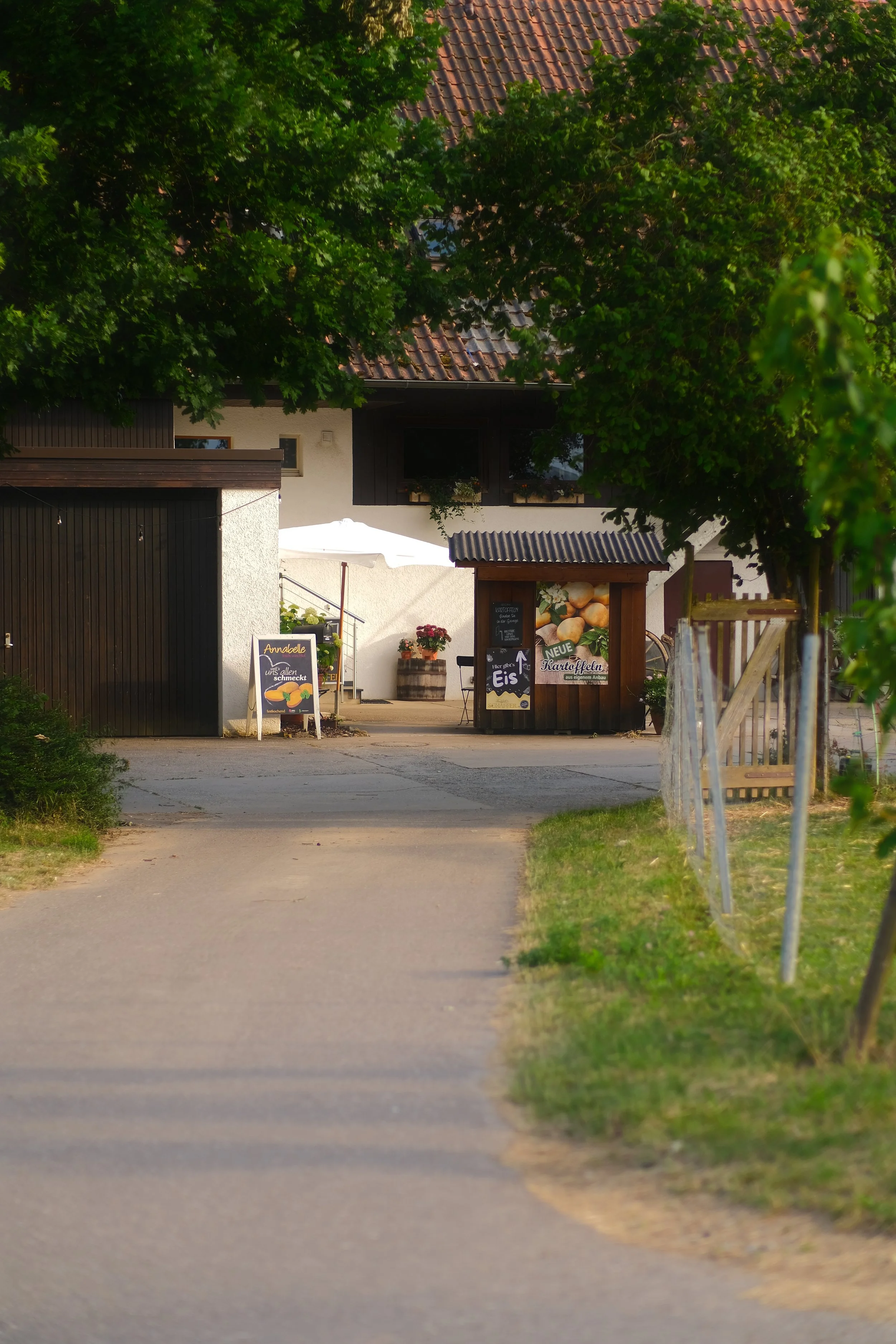Ein schmaler Gehweg führt zu einem Landgasthof oder Restaurant, der im Hintergrund liegt, umgeben von Grünflächen, mit einem weißen Sonnenschirm und einem Wegweiser. Es gibt sowohl Holz- als auch Putzfassaden, und im Vordergrund sind Bäume und ein Za