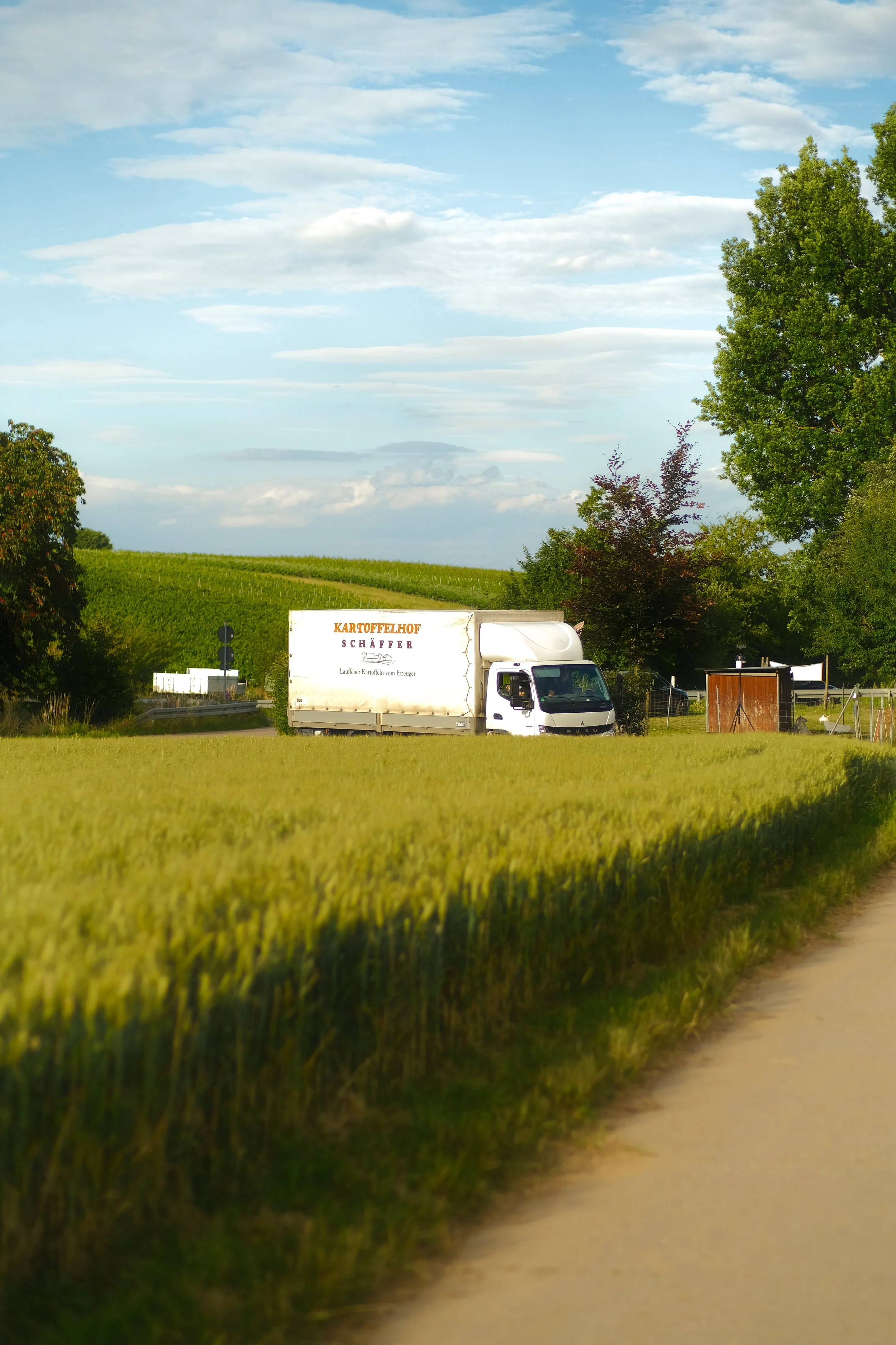Ein weißer LKW mit der Aufschrift 'KARTOFFELHOF SCHÄFFER' parkt am Rand des Feldes, umgeben von Bäumen unter einem blauen Himmel mit Wolken. Das Bild soll den LKW beim Ausliefern bei Wiederverkäufern zeigen.