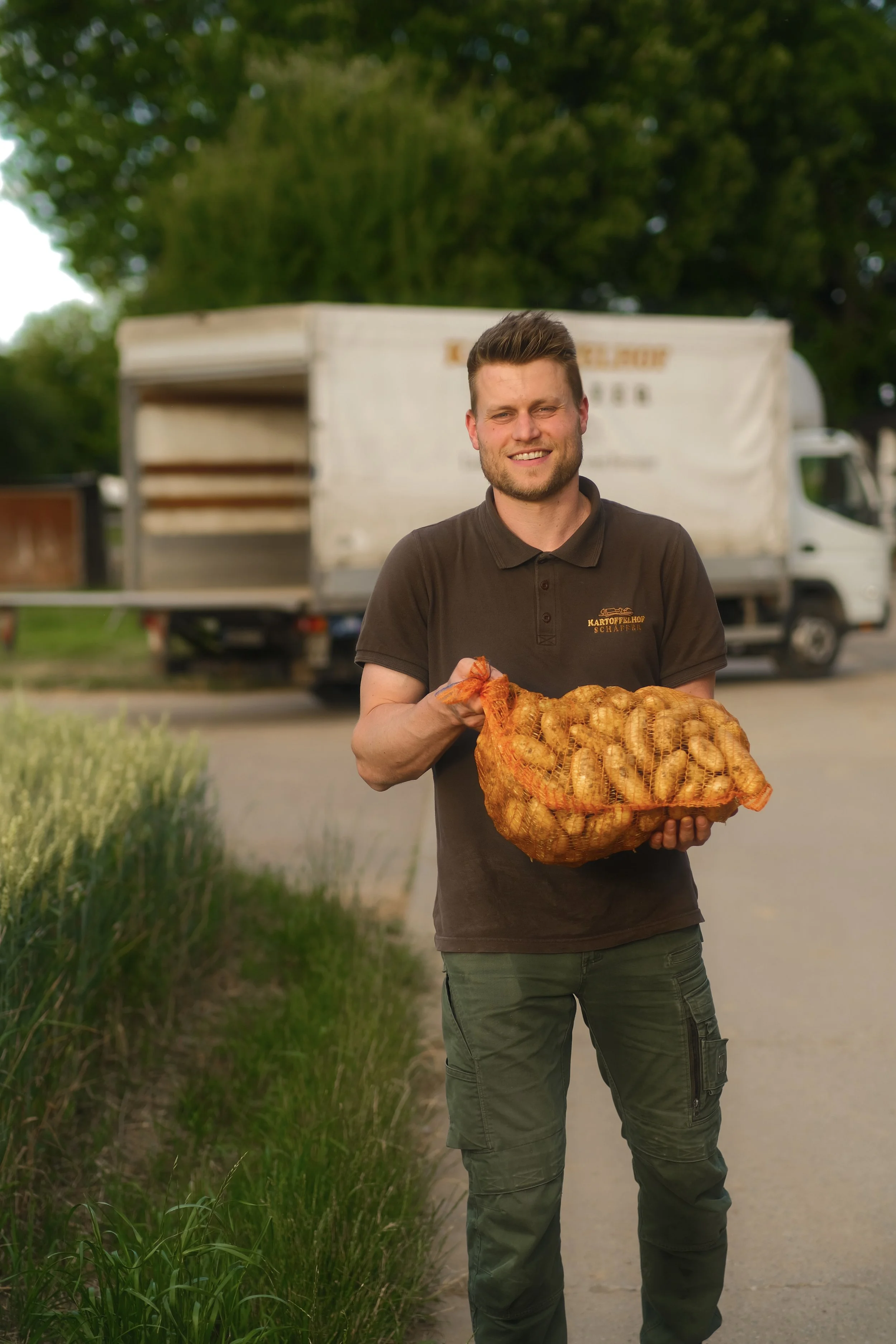 Jonas Schäffer vom Kartoffelhof Schäffer trägt einen großen Korb mit Kartoffeln auf einem Feldweg, im Hintergrund ein Lastwagen, umgeben von Bäumen.