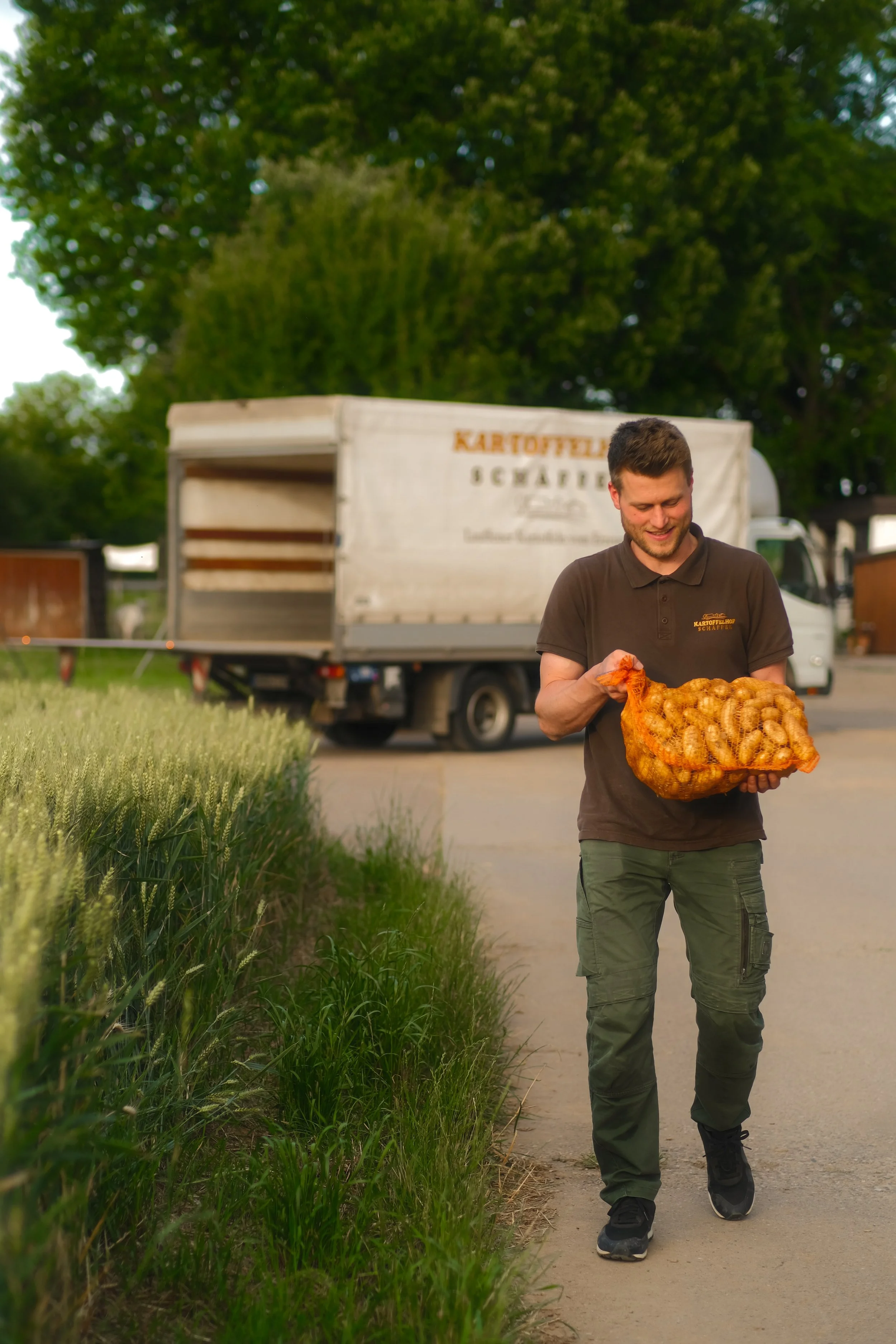Jonas Schäffer vom Kartoffelhof Schäffer trägt einen großen Korb mit Kartoffeln auf einem Feldweg, im Hintergrund ein Lastwagen, umgeben von Bäumen.