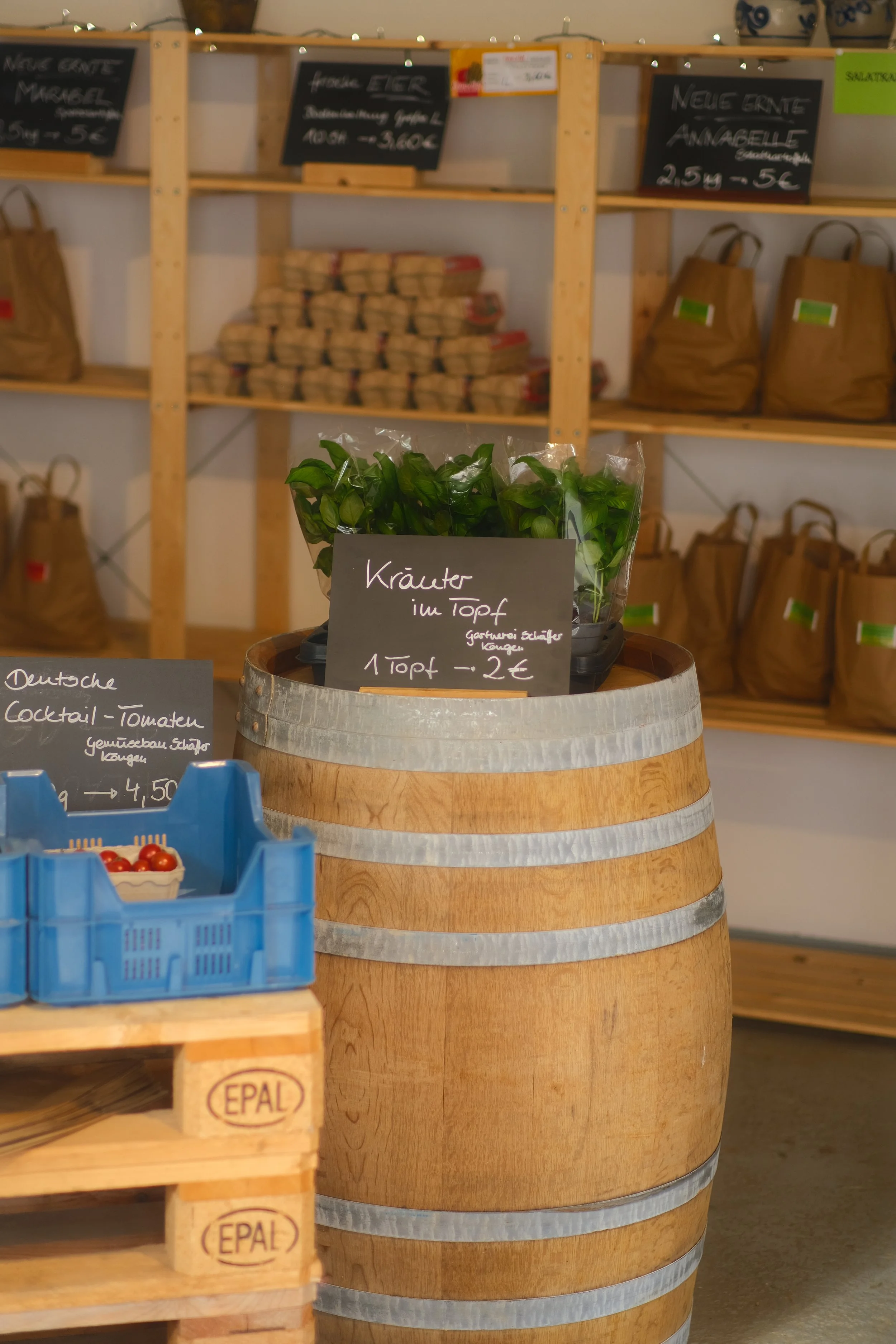 Frische Kräuter in einem kleinen Topf auf einem Bierfass, im Hintergrund Holzwandregale mit verschiedenen Produkten und Schildern.