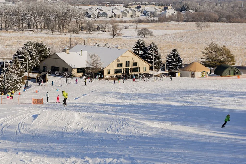 People skiing and snowboarding on a snow-covered slope with a lodge and trees in the background.