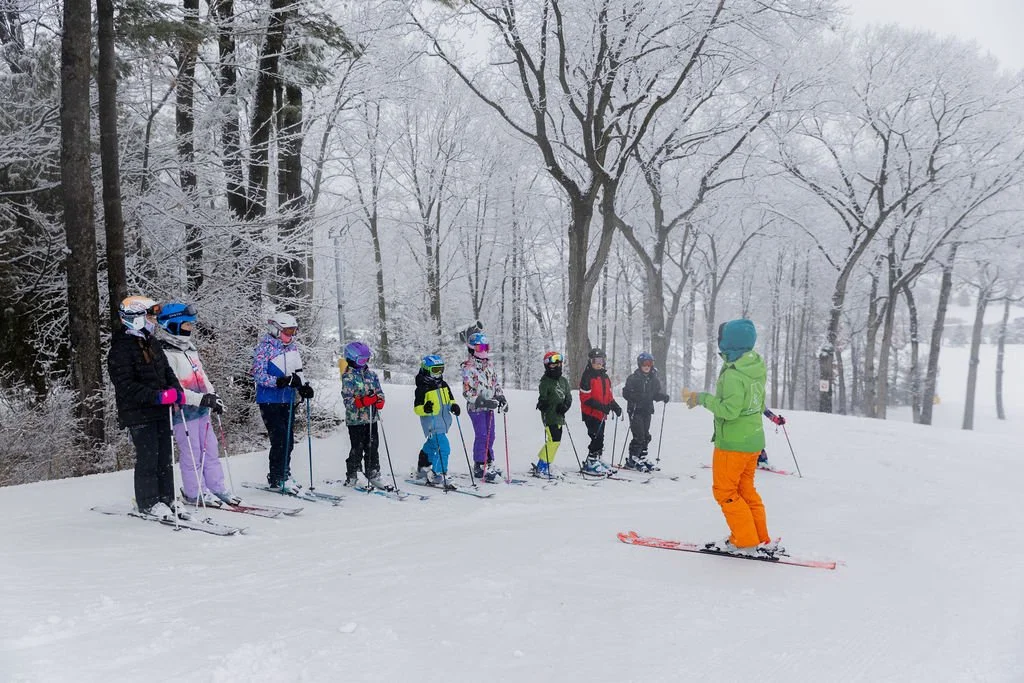 A group of children on skis listening to an instructor in a snowy, wooded area.