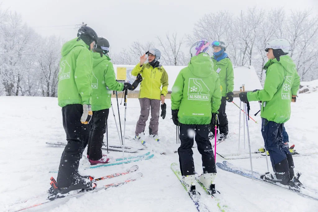 Group of skiers in green jackets listening to an instructor on a snowy mountain.