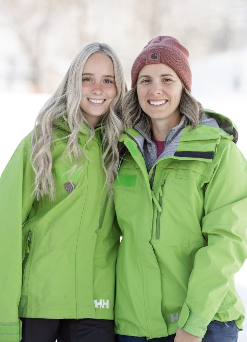 Two women outdoors in winter, both wearing bright green jackets. One woman has long blonde hair, and the other has shoulder-length gray hair and is wearing a red beanie. They are smiling and standing close together.