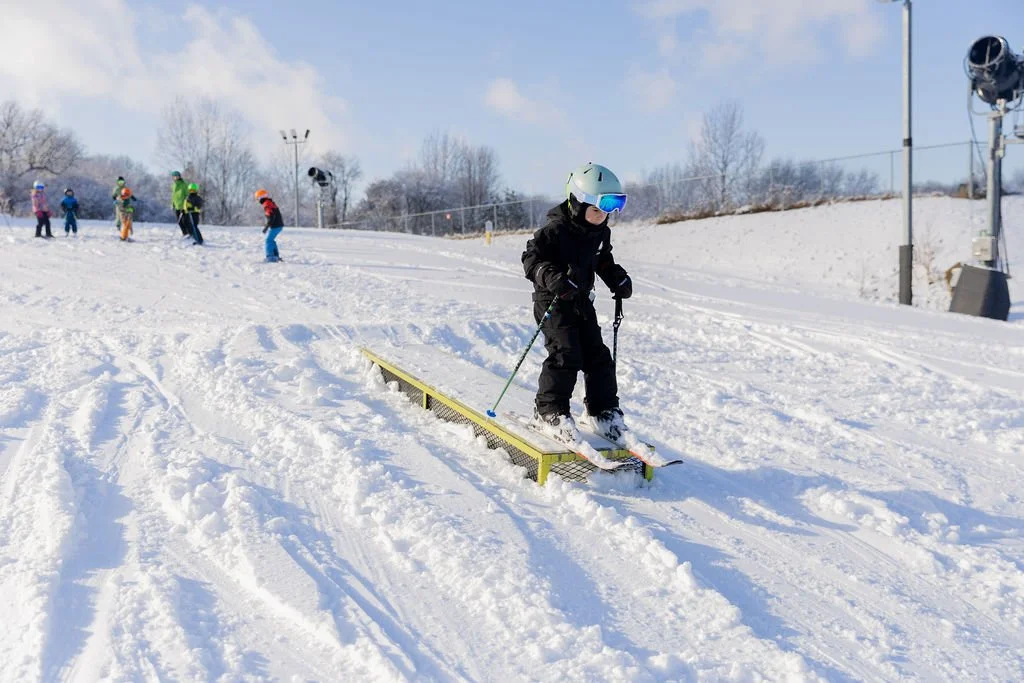 A child in winter gear, including a helmet and goggles, learning to ski on a snow-covered slope with a balance rail, with other children and adults skiing in the background.