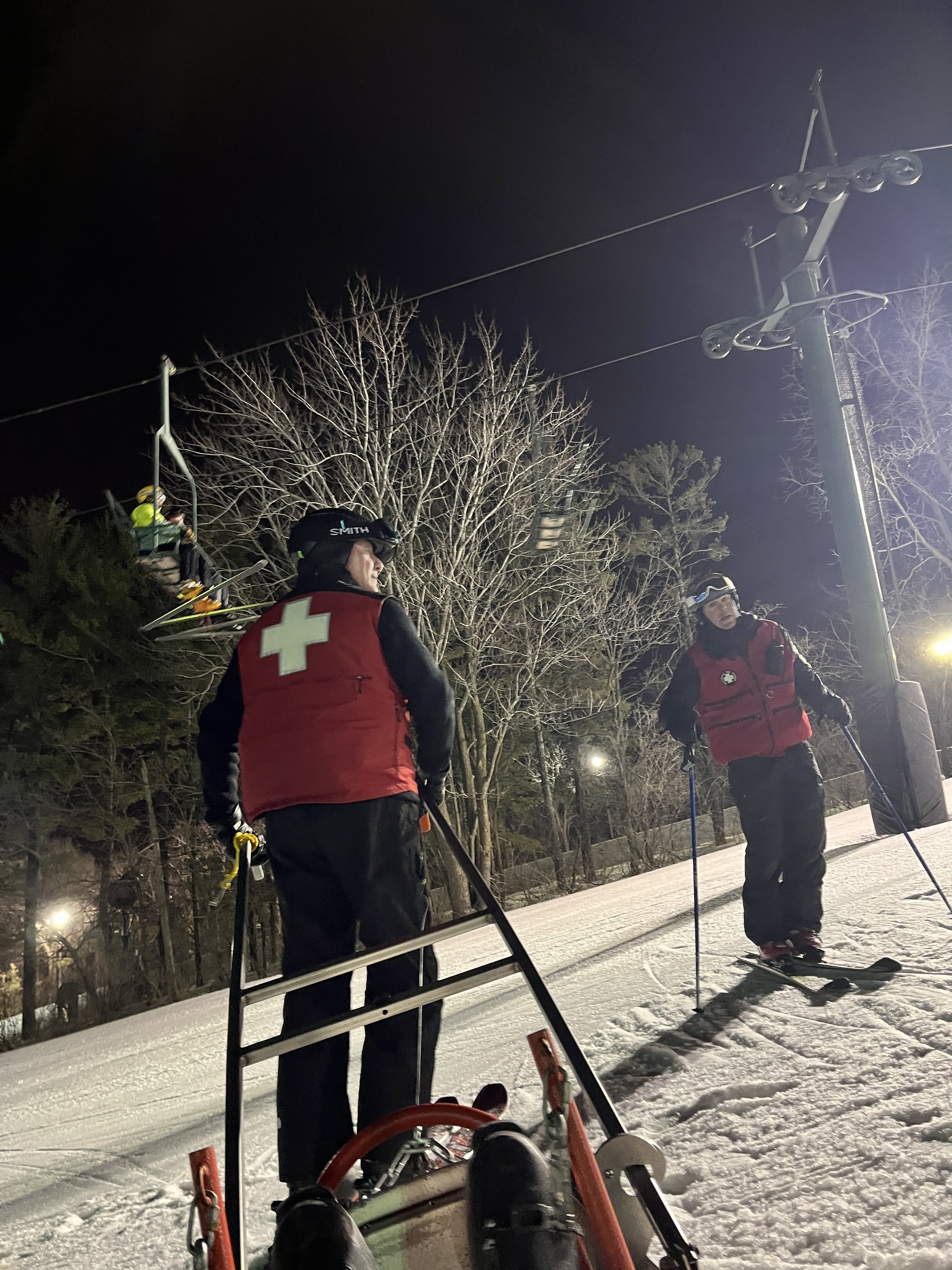 Nighttime scene of three ski patrol members in red vests with medical symbols, standing on snow-covered ground, with ski equipment, and a ski lift structure with large wheels, trees in the background, and dark sky overhead.