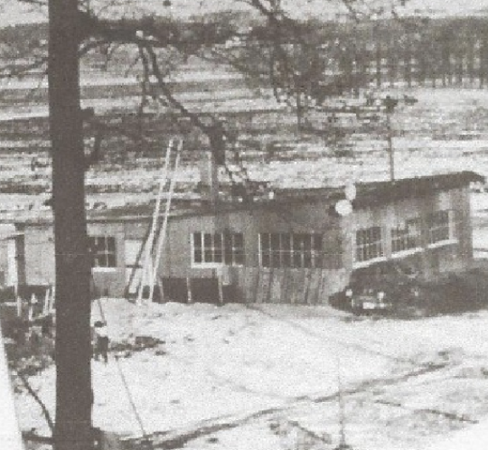 A black and white photo of a small, possibly abandoned building surrounded by snow and trees.