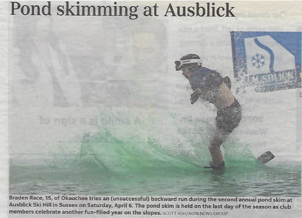 A person wearing a helmet and black shorts stands in a small greenish pond at Ausblick Ski Area, attempting a backward ski trick during a pond skim event.
