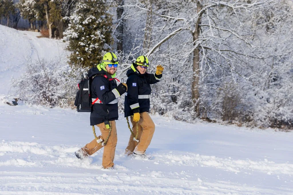 Two rescue workers in cold-weather gear walking in snow, with snow-covered trees in the background.