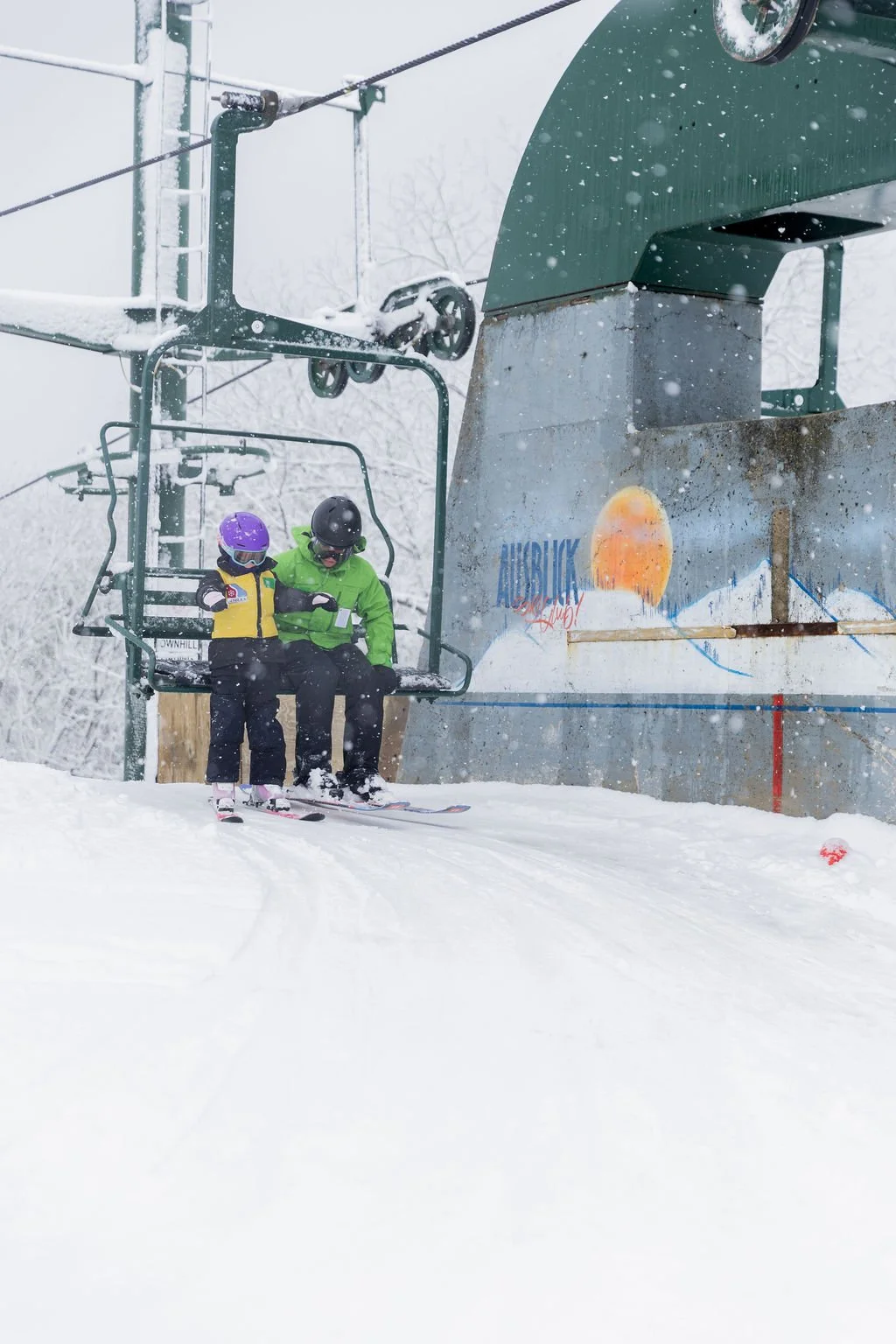 Two skiers sitting on a ski lift, wearing helmets and winter gear, with snow falling and a mural of a sunset over mountains in the background.