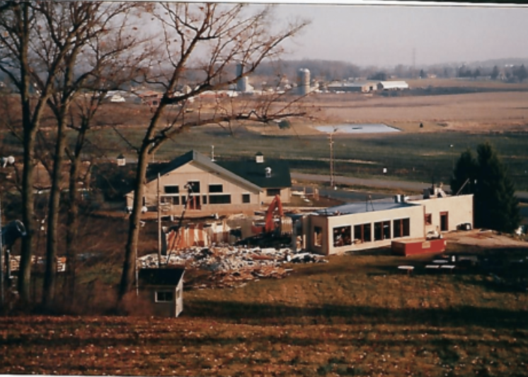 Construction site with a partially built white building, an excavator, and debris, in a rural area with trees and distant barns or silos.