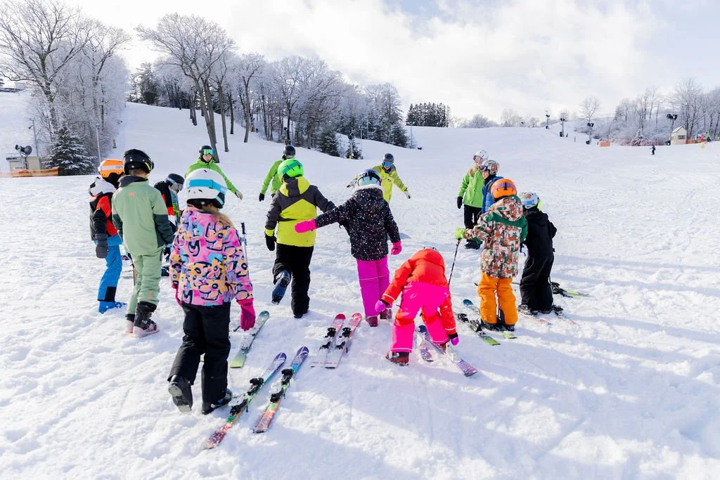 Children in colorful winter clothing and helmets learning to ski on a snowy slope with instructors, surrounded by snow-covered trees.