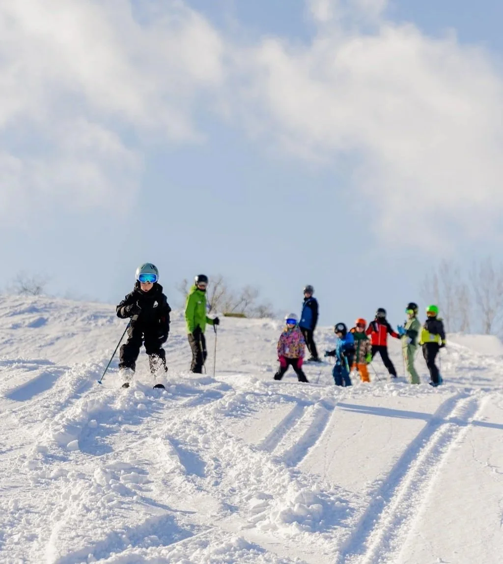 Children and adults in winter clothing skiing and playing on a snowy hill outdoors under a partly cloudy sky.
