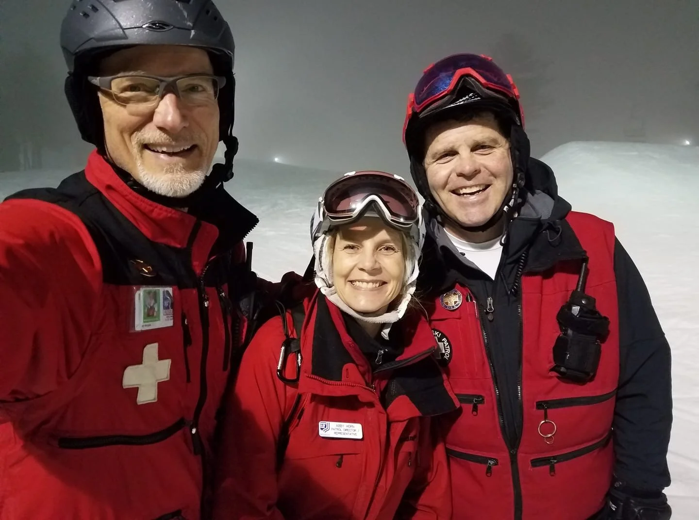 Three smiling rescue workers in red and black jackets with helmets and goggles, standing in a snowy environment.