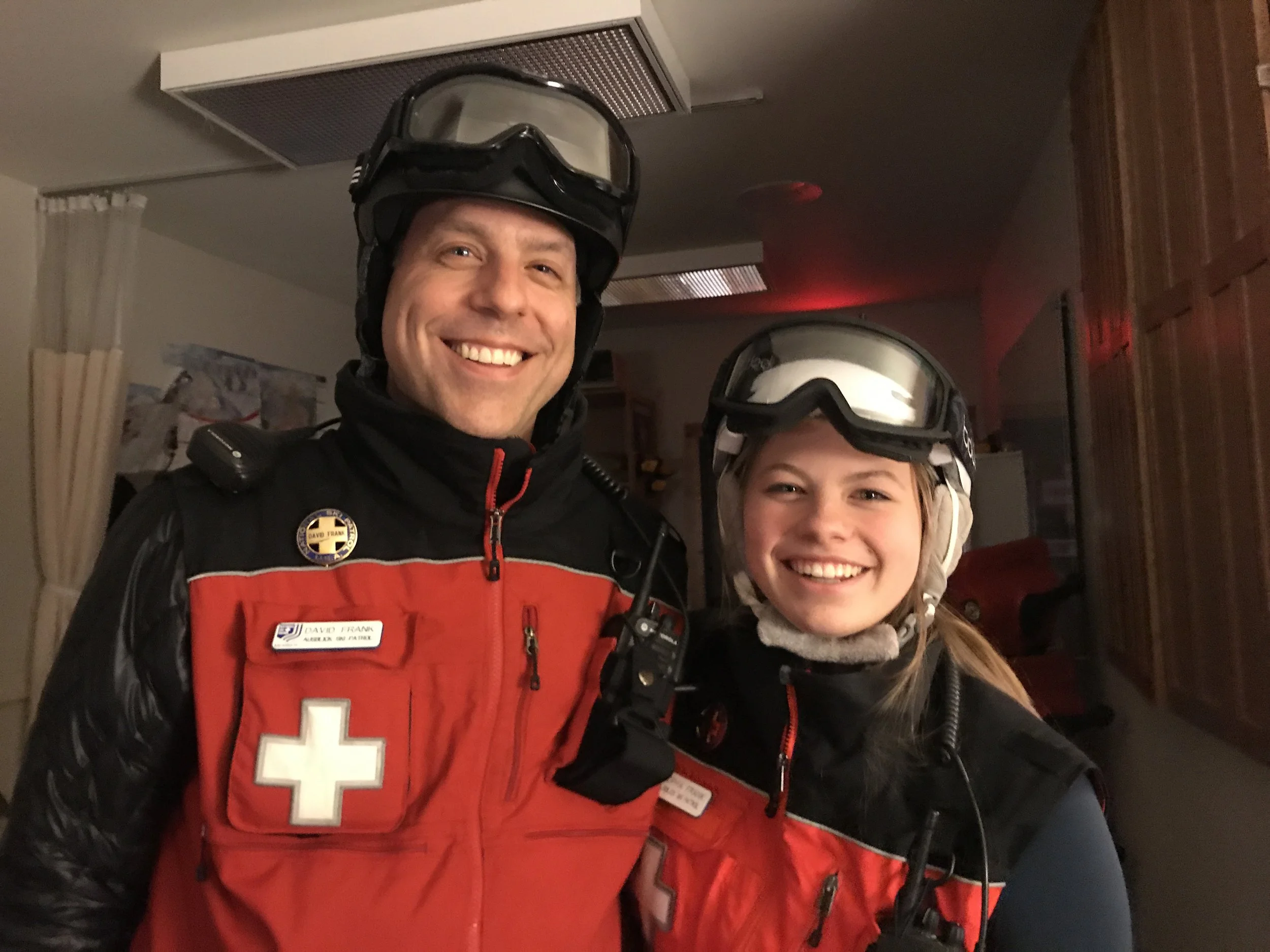 A smiling man wearing a black helmet and a red emergency medical vest with a white cross and name tag, standing indoors with a young girl also wearing a helmet.
