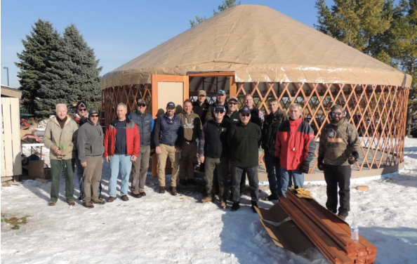 A group of people standing outside in front of a partially constructed yurt on snowy ground, with a clear blue sky and trees in the background.
