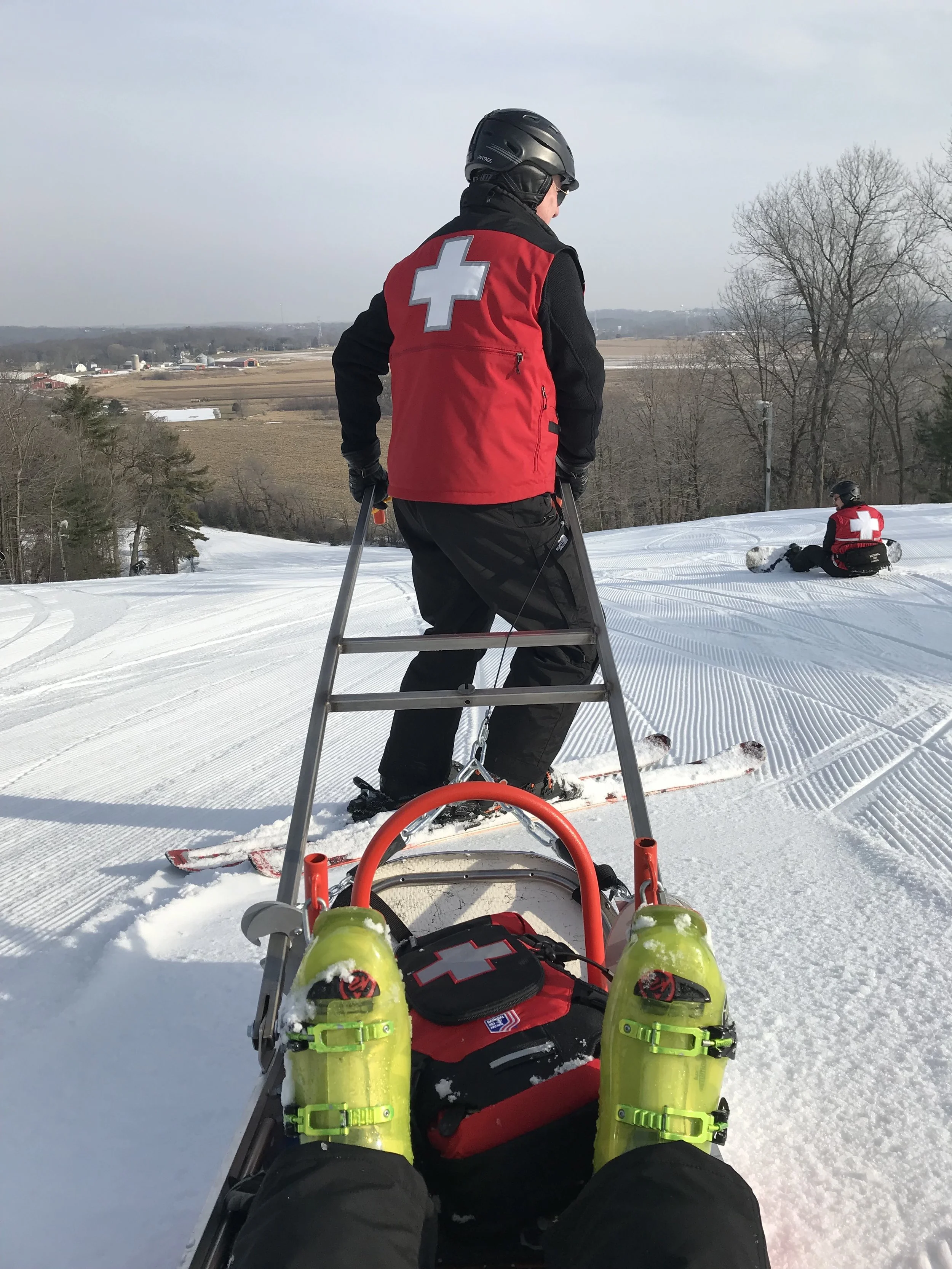 Person on a snow-covered trail wearing a red vest with a white cross, helmet, and sunglasses, looking at the horizon, with another person in similar attire sitting in the snow in the background.