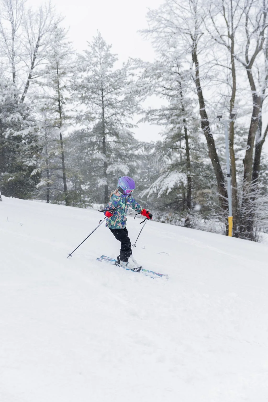 A young girl skiing down a snowy slope wearing a colorful winter jacket, purple helmet, and black pants, with snow-covered trees in the background.