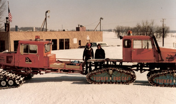 A snowcat vehicle with two people standing on it, parked on snow in front of a small commercial building, with power lines and trees in the background.