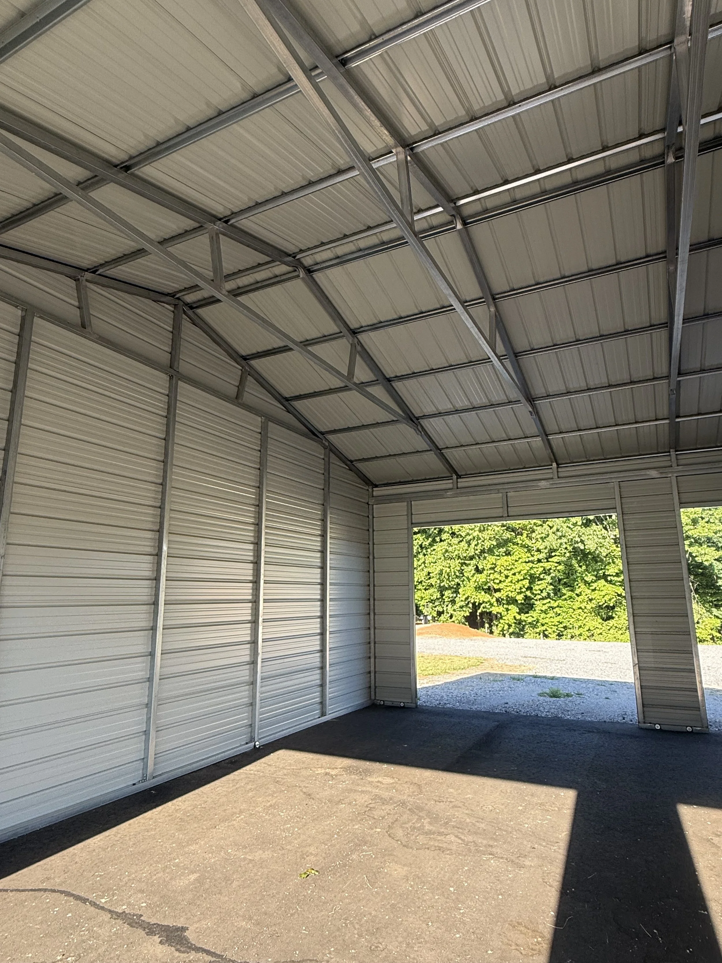 Inside view of a metal storage shed with open doors, showing a gravel driveway and green trees outside.